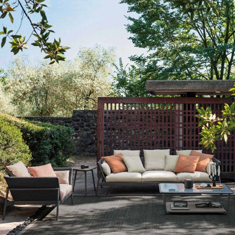 Große Sitzgarnitur mit Sofa und Stuhl stehen mit einem Tisch in einem Garten mit viel Grün im Schatten
