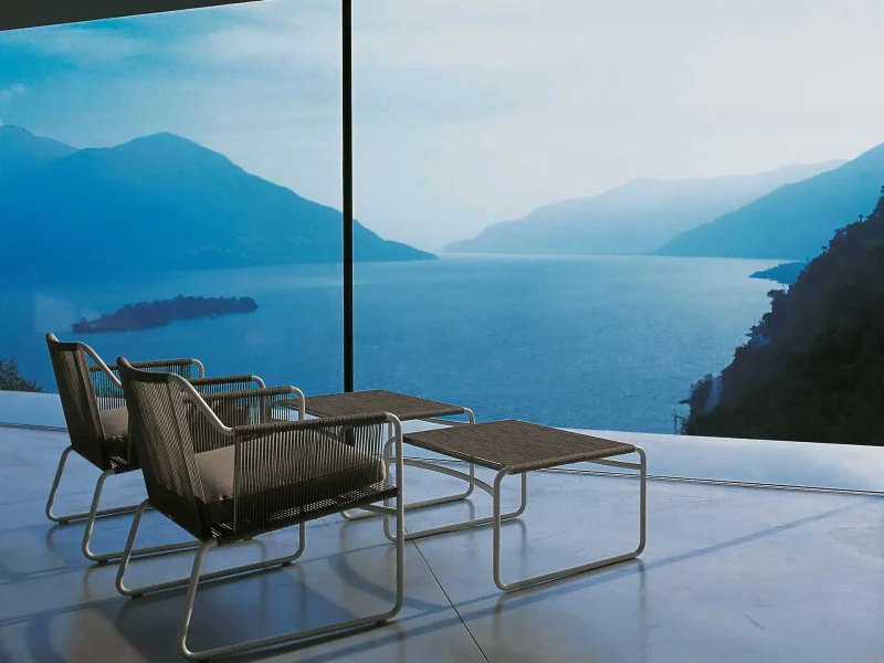 Stühle und Tisch vor einer Glasfront mit Blick auf einen Bergsee.