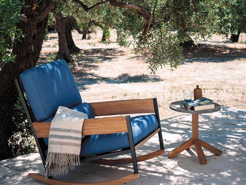 Ein Schaukelstuhl mit blauem Polster steht im Schatten eines Baumes.